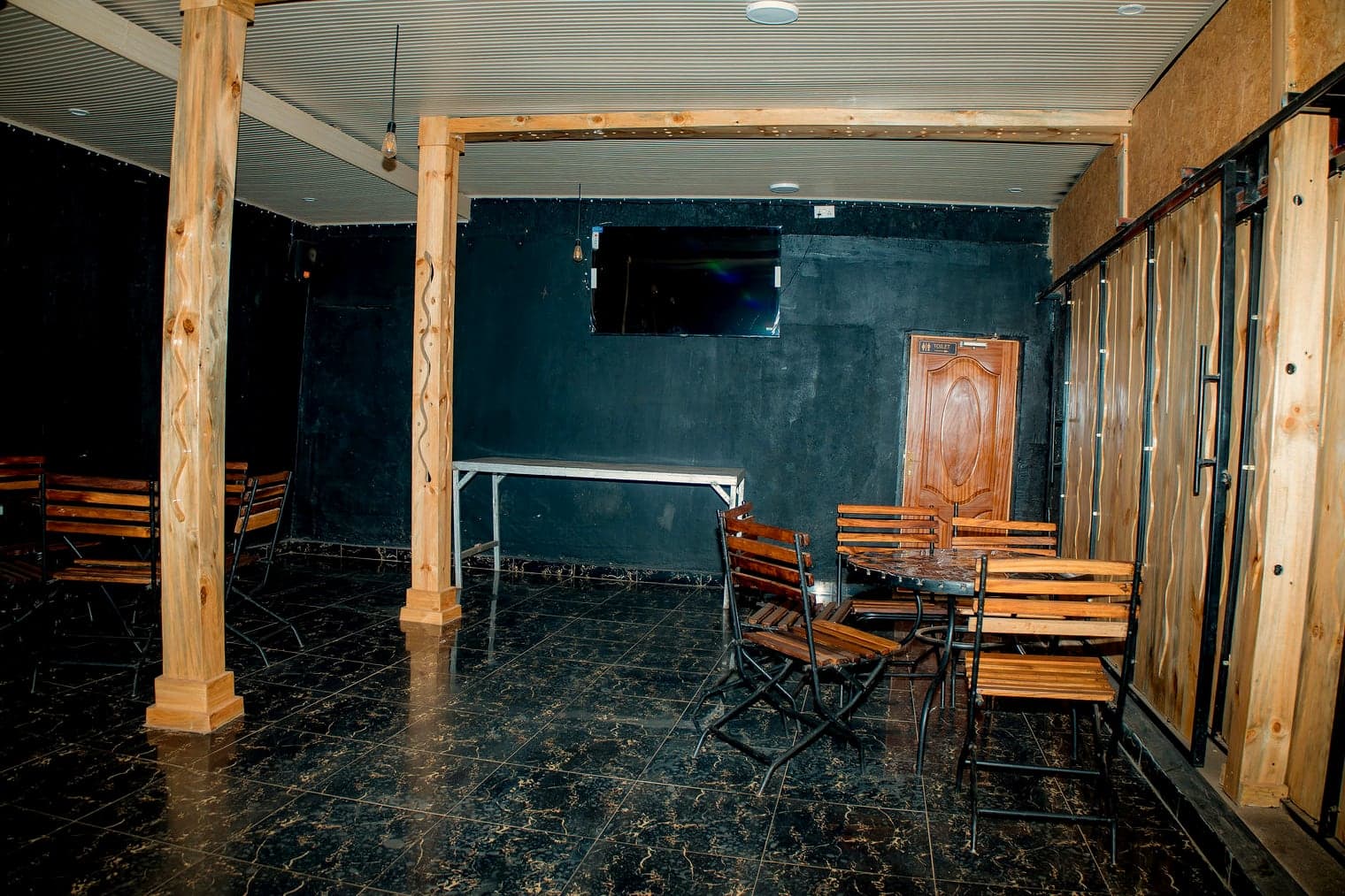 Timber dining chairs and tables under raw wooden beams in the lounge, with black walls and a mounted screen